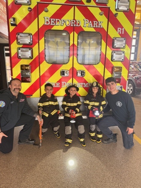 A group of people, including three children in firefighter uniforms, crouch smiling in front of a brightly colored fire truck labeled "Bedford Park."