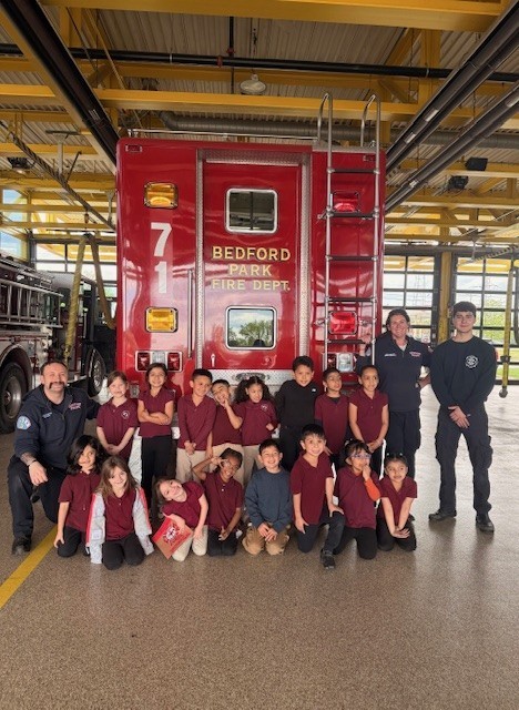 A group of children in maroon shirts and three firefighters pose in front of a red fire truck inside a bright fire station, smiling and cheerful.