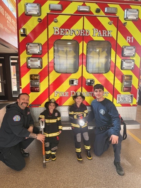 Two firefighters kneel beside two children dressed in firefighter gear, smiling in front of a Bedford Park fire truck. The mood is cheerful and inspiring.