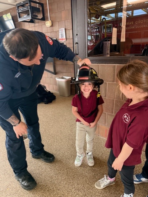 A firefighter puts a large helmet on a smiling child, who wears a maroon shirt, while another child watches. The scene is warm and playful.