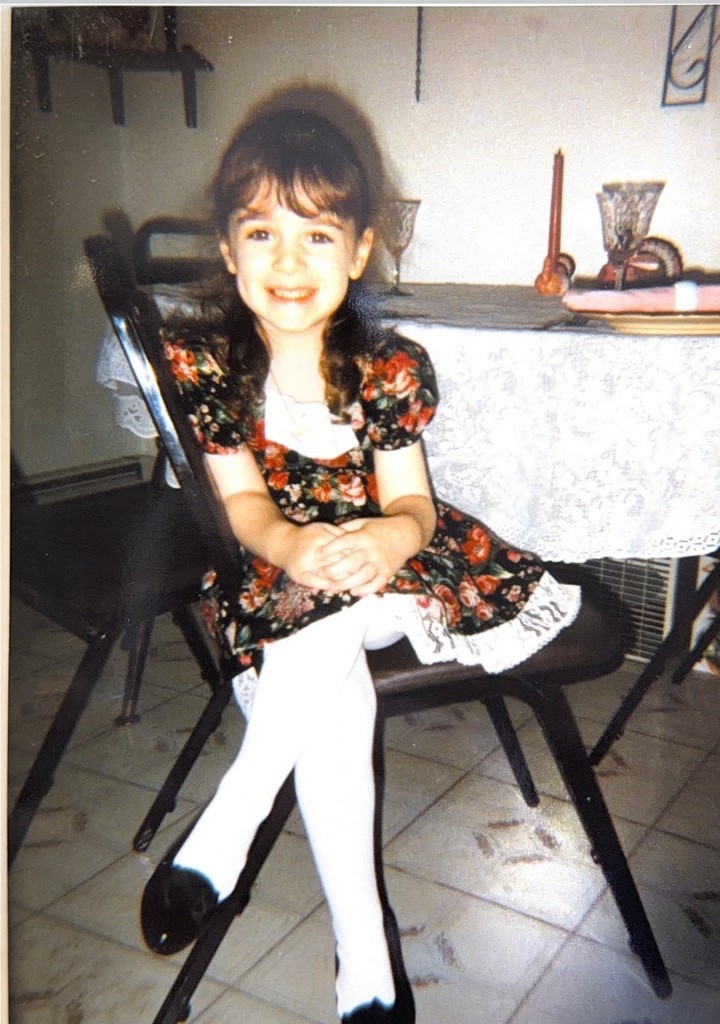 A young girl with braided hair sits on a chair, smiling warmly. She wears a floral dress, white tights, and black shoes. A table with lace cloth and candles is behind her.