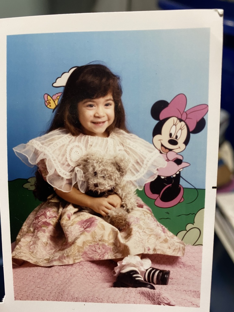 A young girl in a floral dress holds a teddy bear, smiling against a cartoon backdrop with Minnie Mouse. The scene is playful and cheerful.