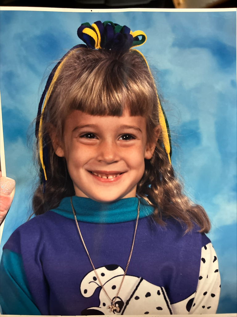 Young girl smiling widely against a blue background. She's wearing a purple shirt with a Dalmatian print, a necklace, and has colorful ribboned hair.