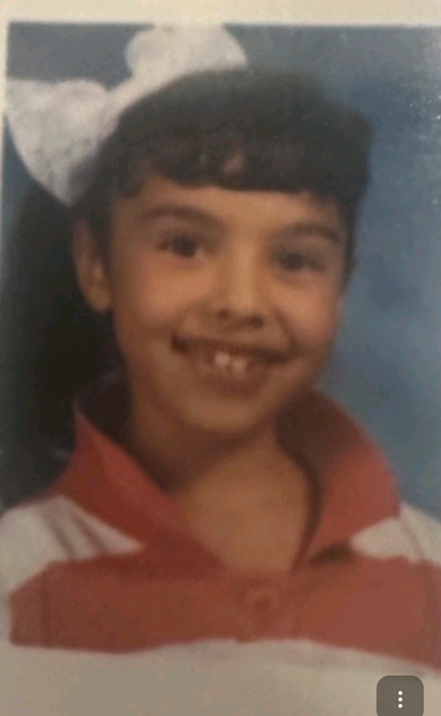 Young child smiling warmly in a school portrait, wearing a red and white striped shirt and a large white bow in their hair against a blue backdrop.