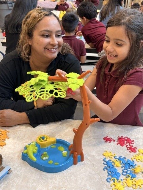 A girl and her teacher happily play with a colorful toy tree at a table. Other children sit in the background. The atmosphere is joyful and playful.