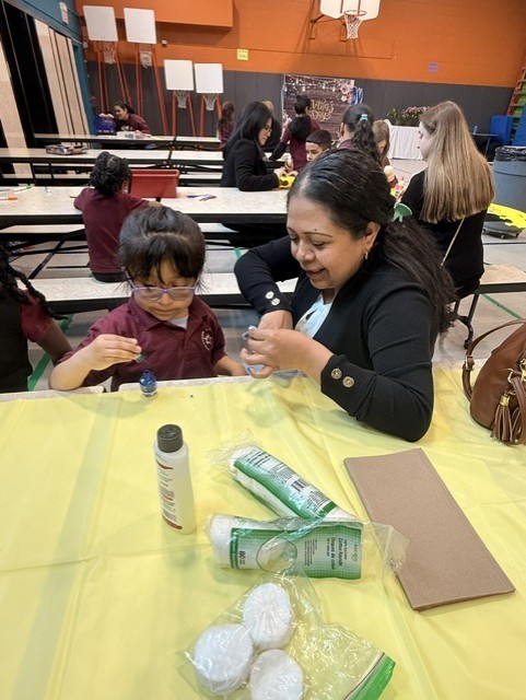 A woman and a young girl are seated at a table with art supplies. The woman smiles, helping the girl with a project. The atmosphere is warm and supportive.