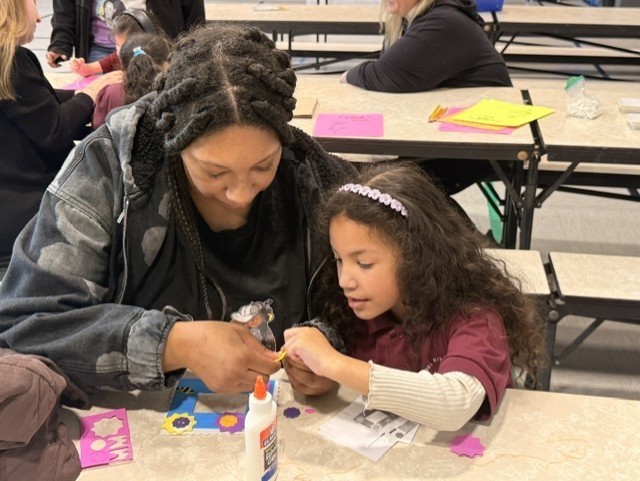 A woman and a young girl are crafting together at a table. They are focused on gluing paper shapes. The scene is warm and collaborative, set in a classroom.