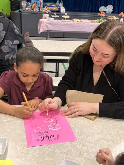 An adult and a child sit at a table, working together on a pink art project with "Best Mom" written on it. Art supplies and snacks are visible in the background.