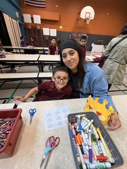 A woman and a smiling child sit together at a table in an art classroom with markers, crayons, and paper. The atmosphere is warm and creative.