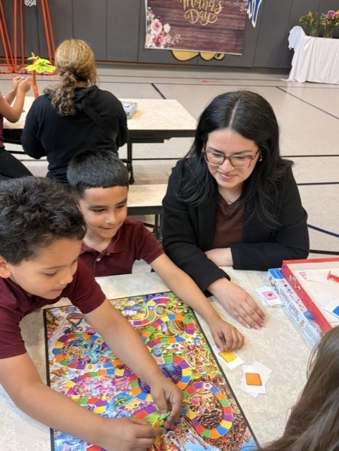 A girl and her teacher happily play with a colorful toy tree at a table. Other children sit in the background. The atmosphere is joyful and playful.