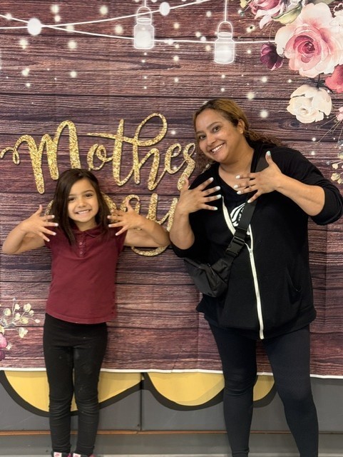 Mother and daughter smiling, posing with jazz hands in front of a floral and wooden backdrop with the words "Mother's Day." The scene is joyful.