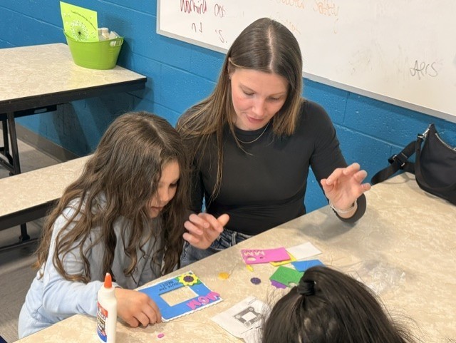 A woman and a girl are seated at a table in a classroom, engaging in a craft activity. They are focused on colorful paper pieces and a glue bottle.