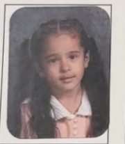 A young child with long dark hair sits against a gray backdrop, wearing a pink top with a white collar. The expression is calm and slightly curious.