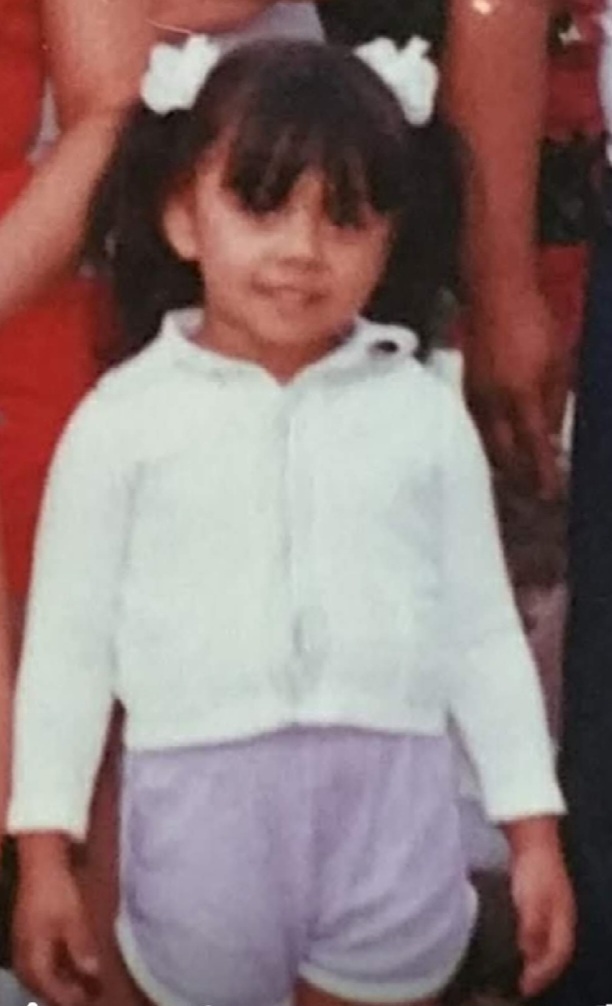 A young girl with pigtails and white bows stands smiling. She wears a white top and lavender shorts. The tone is cheerful and warm, with casual attire.