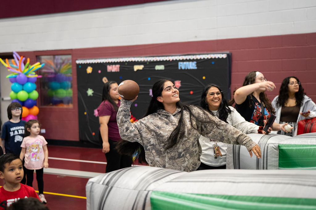 A young woman in a camouflage hoodie joyfully throws a football at an indoor event. Children and adults watch, surrounded by colorful balloons.