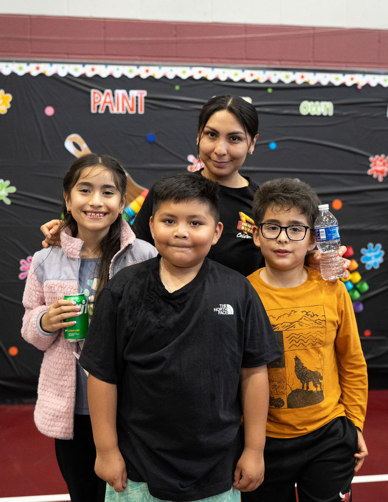 A cheerful group photo shows three children and an adult indoors. The kids smile, holding drinks. A colorful painted banner is in the background.
