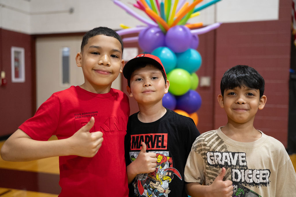 Three boys smiling and giving thumbs up in a gym; behind them, a colorful balloon arrangement. The mood is cheerful and celebratory.
