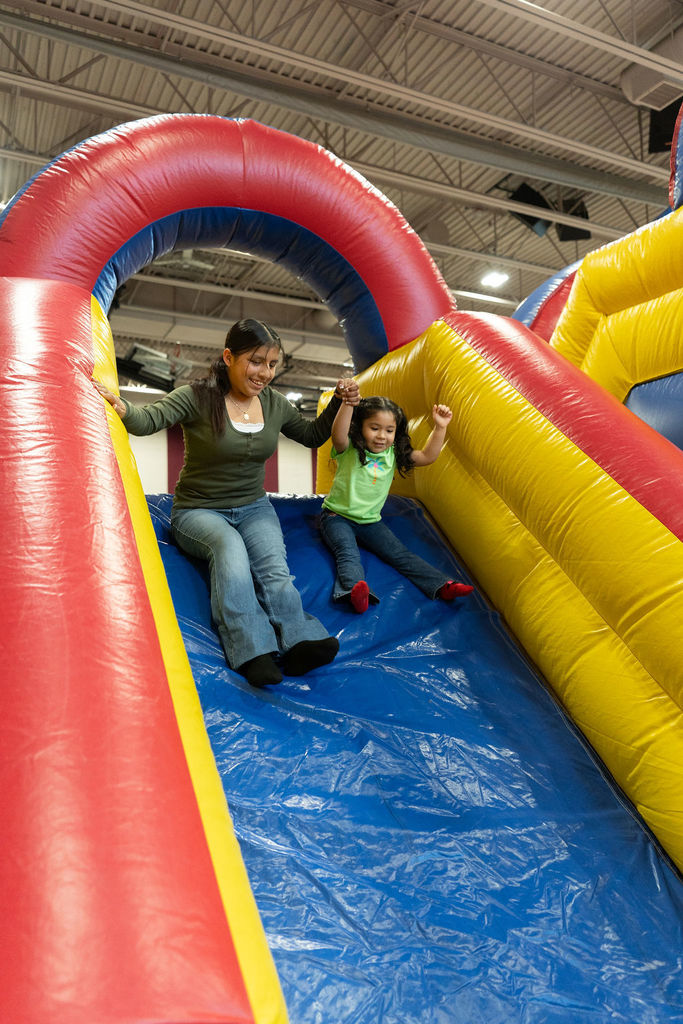 A woman and a young girl joyfully slide down a colorful inflatable slide, holding hands. The setting is indoors, creating a cheerful atmosphere.