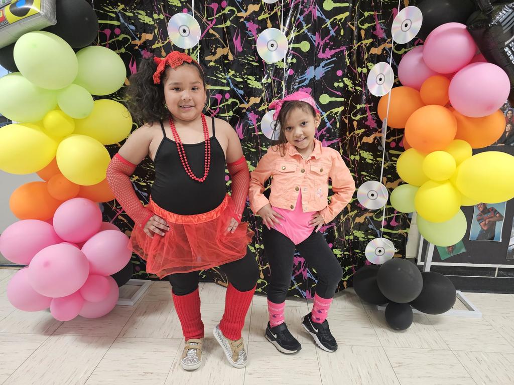 Two smiling children in colorful 80s outfits pose in front of a vibrant, splatter-painted backdrop with balloons and CDs, exuding joy and playfulness.