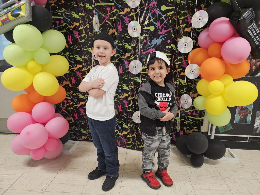 Two young boys pose confidently in front of a colorful 80s-themed backdrop with neon paint splatters and CDs. They are flanked by vibrant balloon arches.