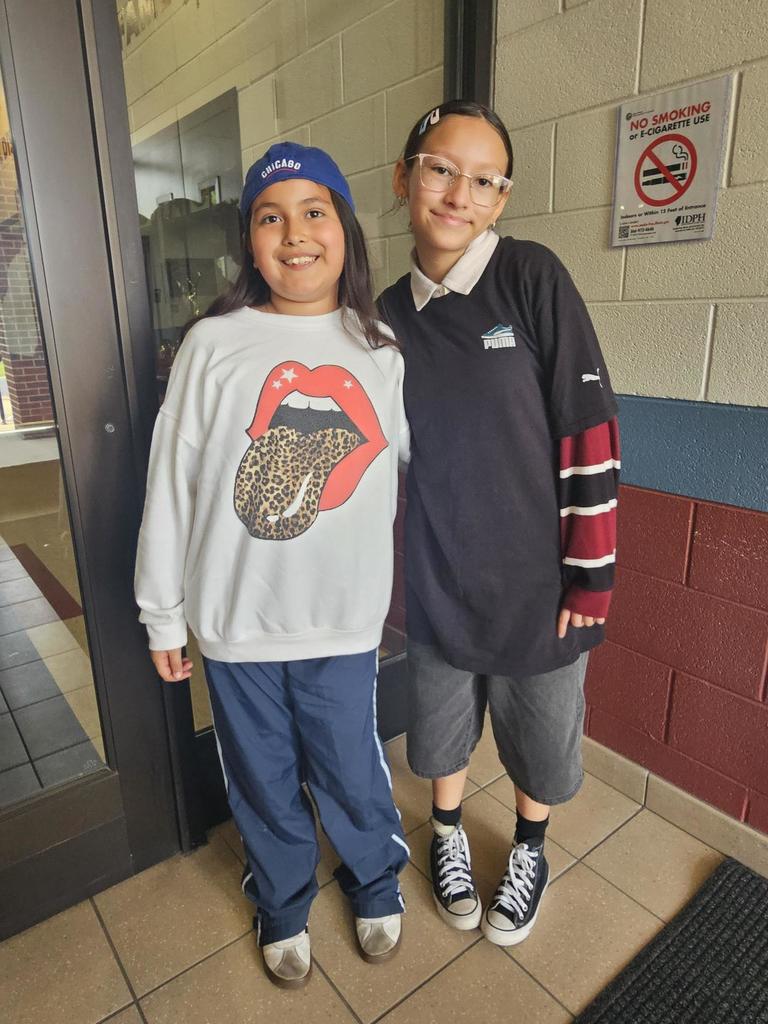Two smiling children stand side by side indoors. One wears a graphic sweatshirt and blue cap, the other in a black shirt with striped sleeves. Casual and cheerful mood.