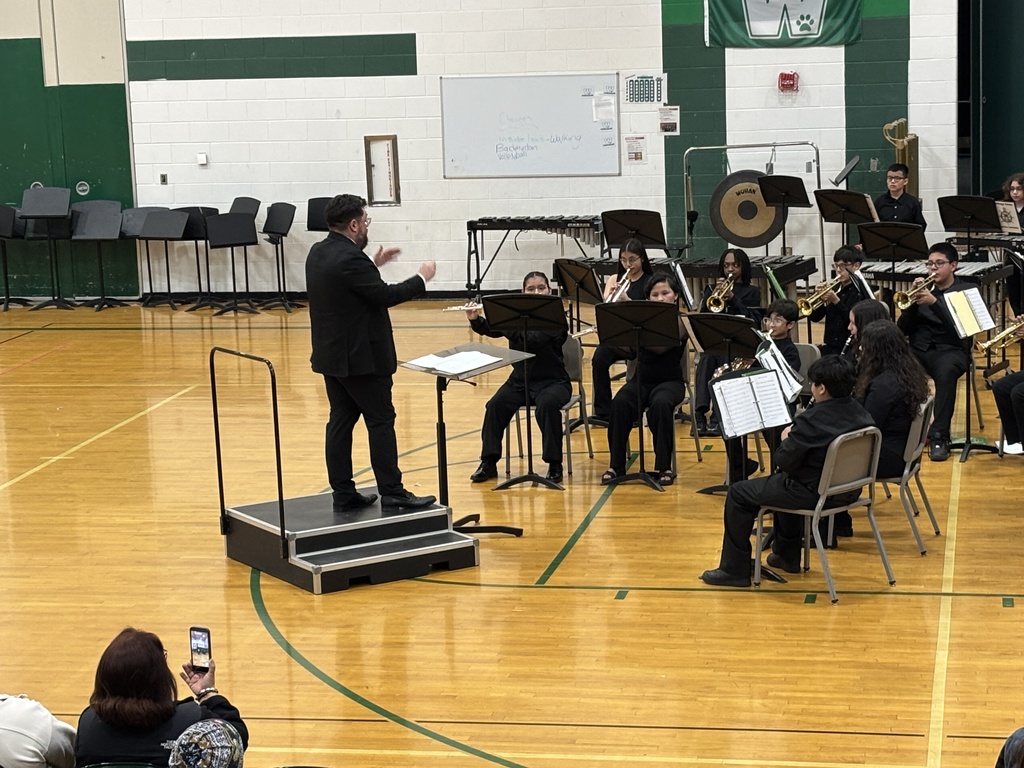 A conductor leads a school band in a gymnasium, with students playing brass and woodwind instruments. Audience members watch, one holds a phone.