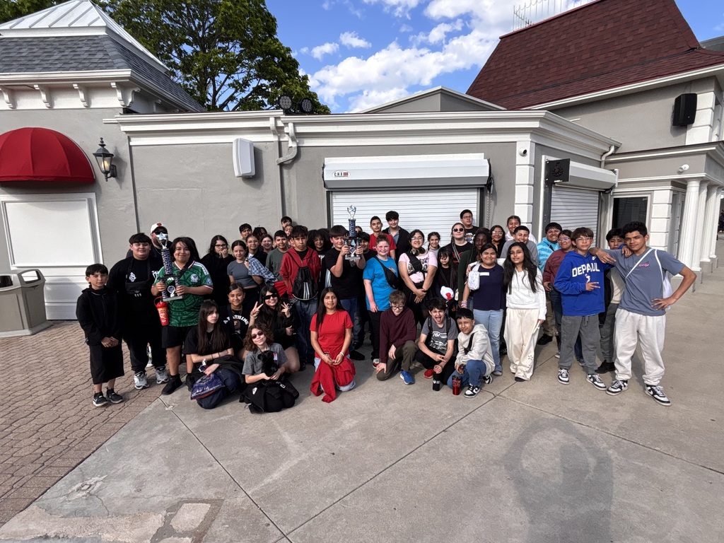 A large group of smiling people stands together outside a building with gray walls and red awnings. The sky is clear and blue, suggesting a joyful atmosphere.