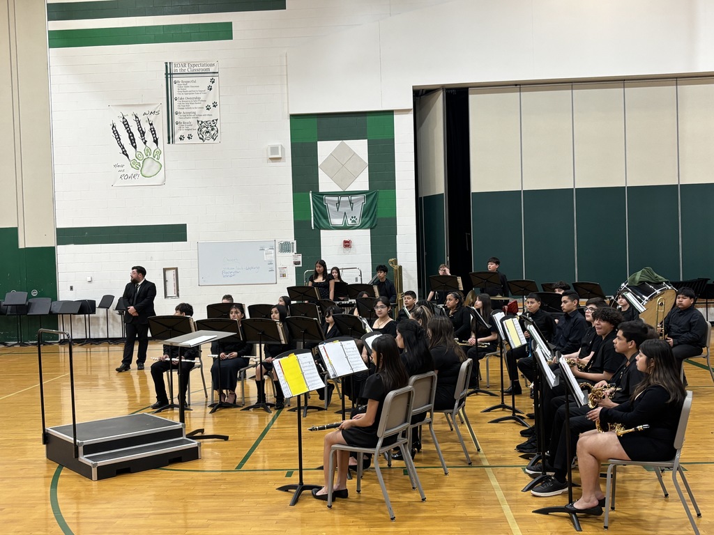 Students in formal attire sit with musical instruments in a green and white gymnasium. A teacher stands beside them, suggesting a concert setting.