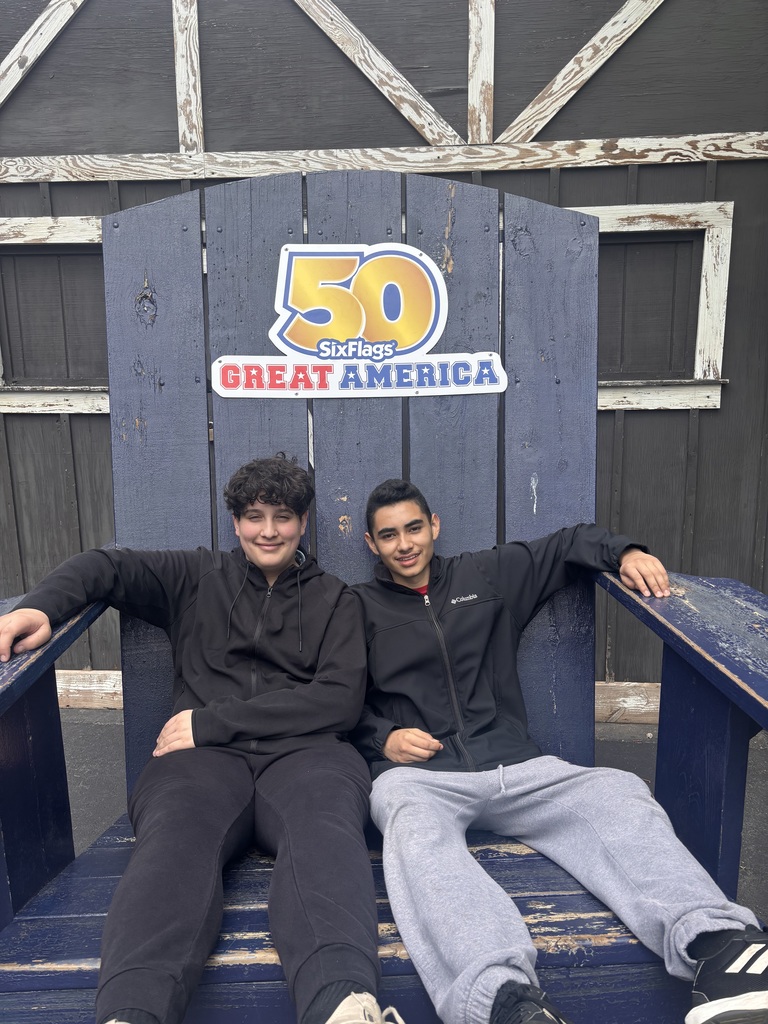 Two smiling individuals sit on a giant blue wooden chair at Six Flags Great America, with a "50" anniversary sign above them. Casual and fun atmosphere.