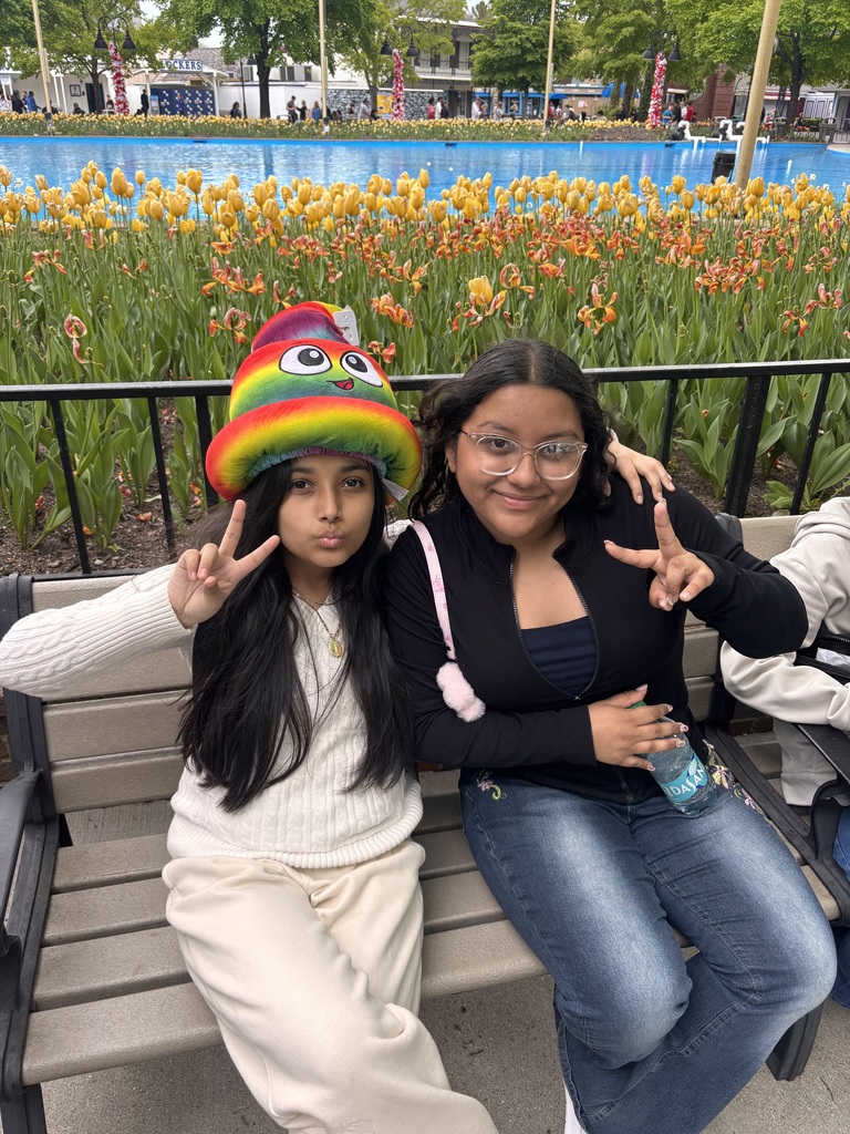 Two people sit on a bench, flashing peace signs. The person on the left wears a colorful rainbow hat, and behind them are vibrant orange tulips and a blue fountain. The mood is playful and cheerful.