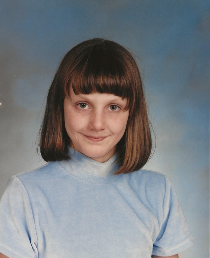 A young girl with straight brown hair and bangs smiles softly in a school portrait. She wears a light blue turtleneck against a blue backdrop.