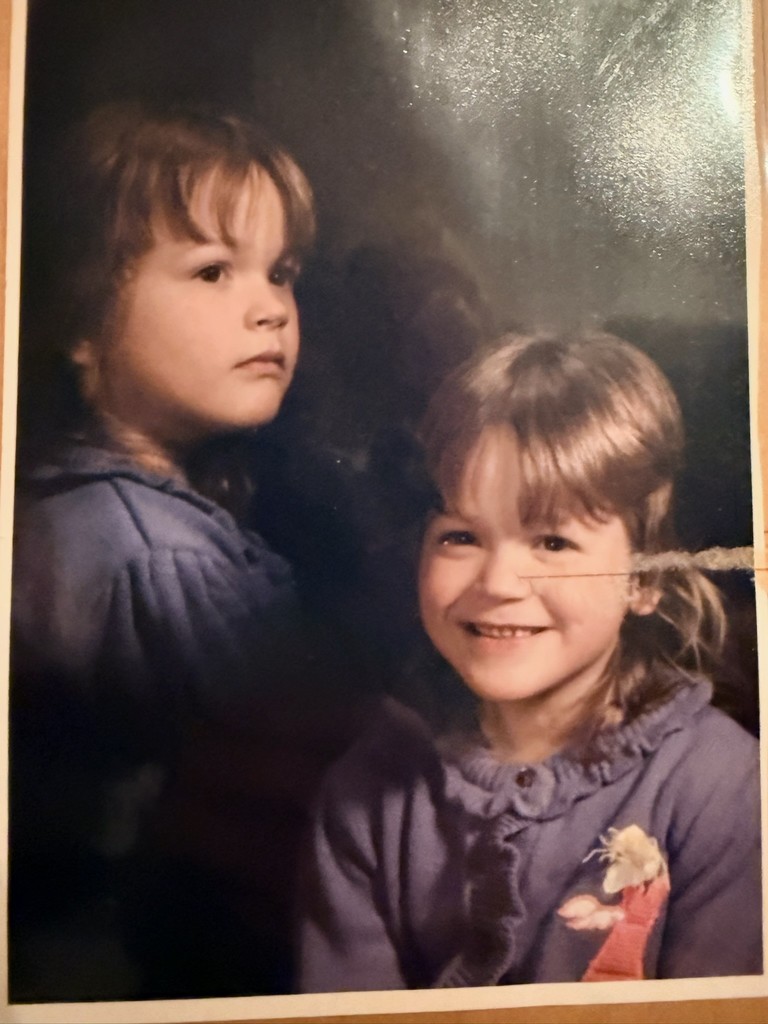 Two young children are depicted, one smiling at the camera and the other gazing sideways with a serious expression. Both wear purple sweaters, creating a nostalgic and tender mood.
