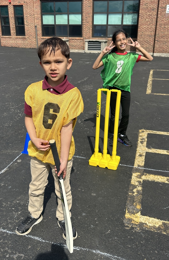 Two children are playing cricket on a playground. The boy in front holds a bat, wearing a yellow vest, while the girl behind poses happily in a green vest.