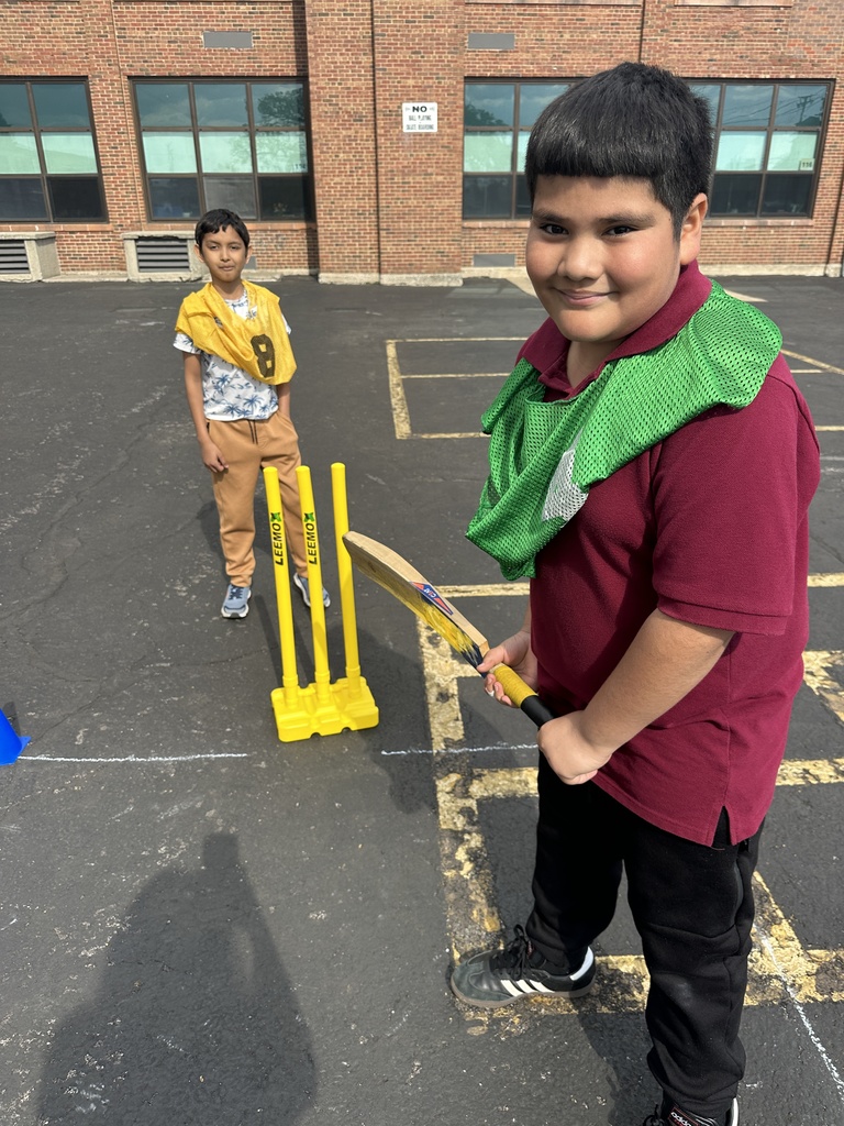 Two boys playing cricket on a paved area. One in a maroon shirt holds a bat with a green bib, smiling. The other stands by yellow stumps wearing a yellow bib.