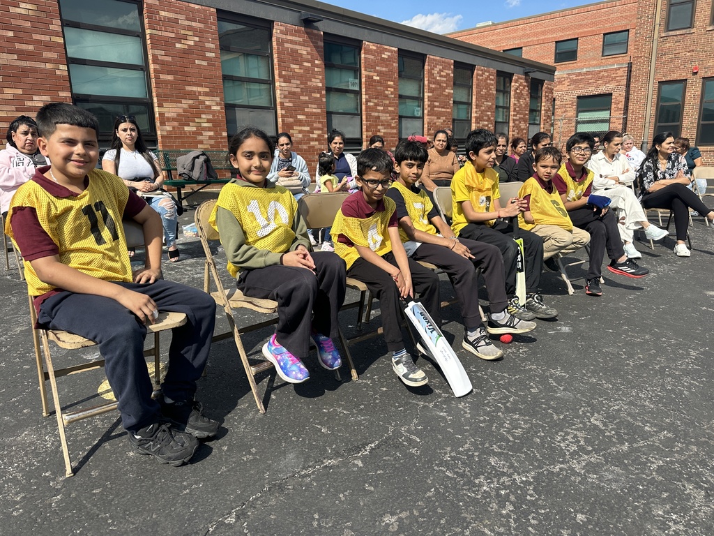 Children in yellow sports jerseys sit in a row on folding chairs, holding cricket equipment, with adults seated in the background. The mood is cheerful.