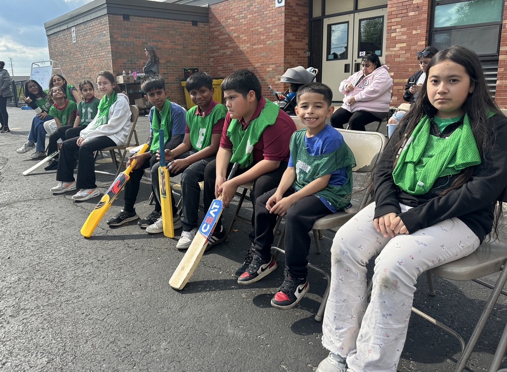 A group of children wearing green sports vests sit in a row on folding chairs outdoors, holding cricket bats. They appear cheerful and engaged.