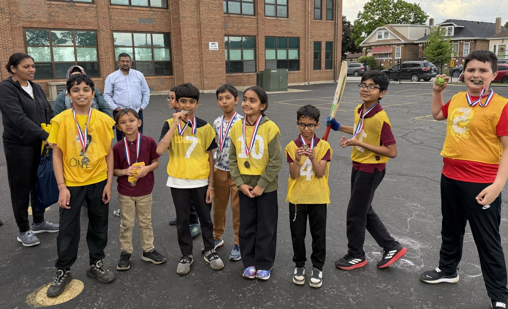 A group of smiling children wearing yellow sports jerseys with medals around their necks stands outside a school building, exuding excitement and pride.
