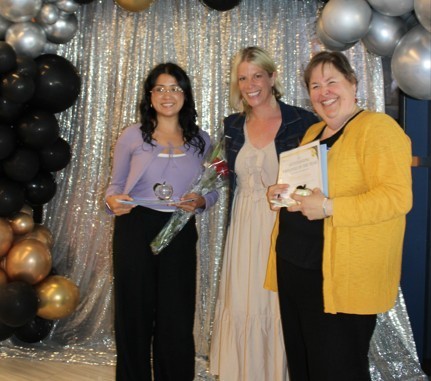 Three women standing in front of a silver, sparkling backdrop with black and gold balloons. They are smiling and holding certificates and a rose.