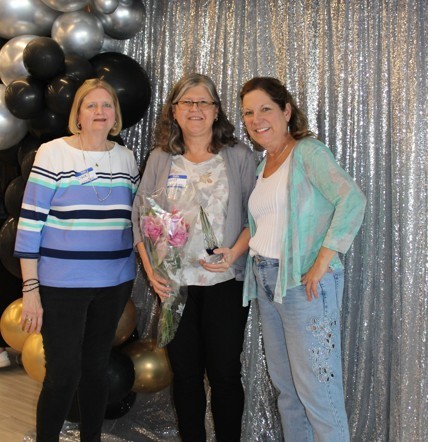 Three women smiling in front of a silver sequin backdrop with black and gold balloons. The woman in the center holds a bouquet of pink flowers, conveying celebration and joy.