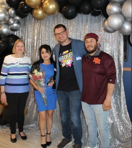 Four people stand smiling in front of silver sequin backdrop and black, gold, and silver balloons. One woman holds a bouquet of red and white roses.