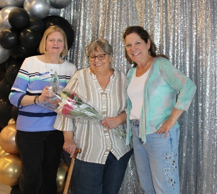 Three smiling women pose in front of a silver sequin backdrop with black and gold balloons. The woman in the middle holds a bouquet of roses and a cane.