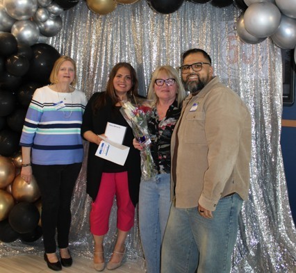 A group of four people smiling in front of a silver sequin backdrop and black, gold, and silver balloons. One woman holds flowers and an envelope.