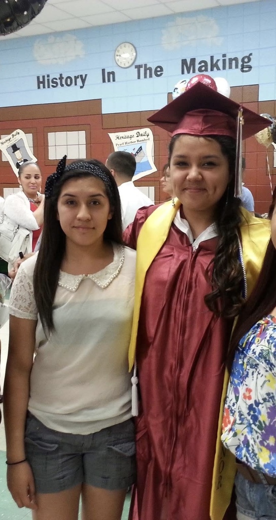 Two young women stand side by side. The one on the right wears a maroon graduation cap and gown, smiling, symbolizing achievement. The background has a wall with the phrase “History in the Making.”