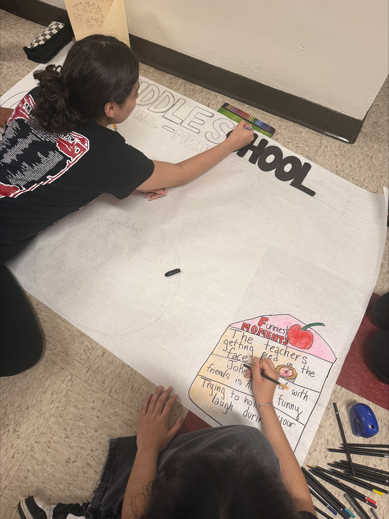 Two students are creating a school poster on the floor. One writes "MIDDLE SCHOOL" and the other adds details to a "Funniest Moments" section with drawings.