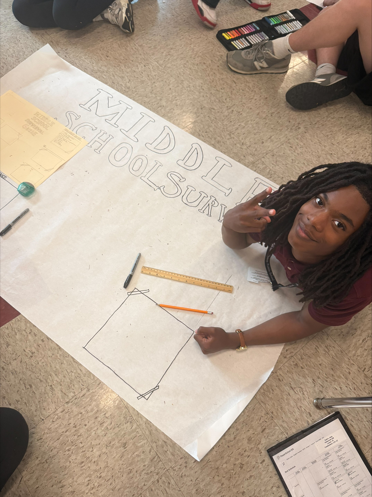 Young student with dreadlocks sits on the floor, smiling, next to a large poster labeled "Middle School Survey," with markers and rulers. Others' legs visible.