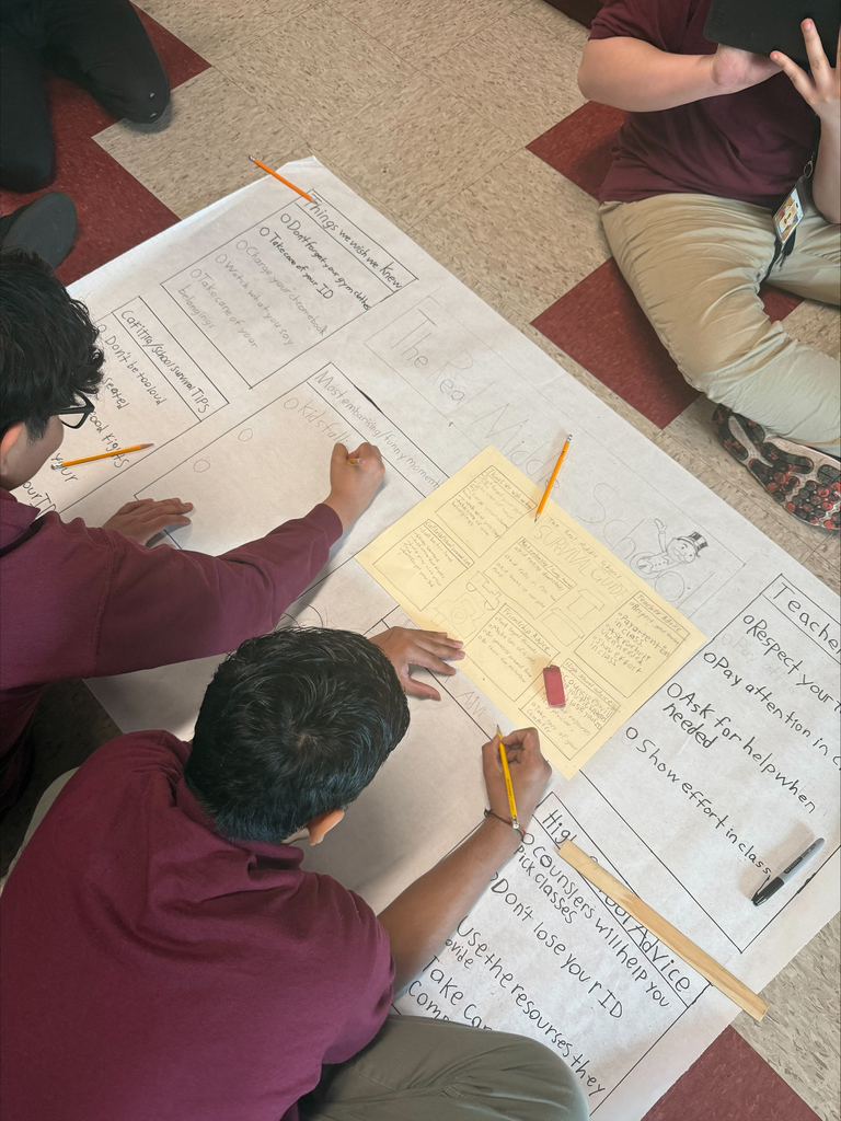 Three students in maroon shirts collaborate on a large poster on a tiled floor. They are writing and sketching with pencils, showing teamwork.