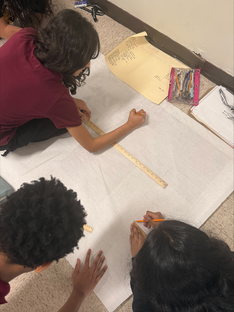 Three students work together on a large sheet of paper, using rulers and pencils. Nearby, notes and art supplies are scattered on the floor.
