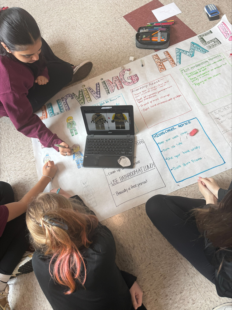 Three students sit on the floor, working on a colorful poster titled "Surviving HM." A laptop with LEGO images is nearby, and art supplies are scattered around.