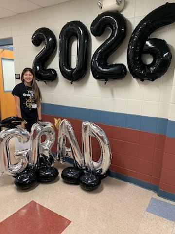 A person stands next to large black and silver balloons spelling "2026 GRAD" in a school hallway, smiling, evoking a celebratory and hopeful tone.