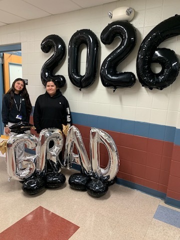 Two people stand in a hallway with large black balloons displaying "2026" and silver balloons spelling "GRAD." The setting conveys a celebratory graduation theme.